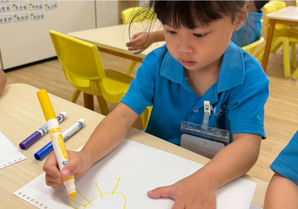 Child in blue shirt draws a yellow sun on paper with a marker at a classroom table.